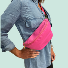 Load image into Gallery viewer, A person wearing a denim shirt is showcasing a bright pink nylon sling bum hip bag with an adjustable strap.