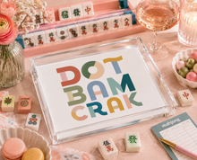 Load image into Gallery viewer, Colorful 'Dot Bam Crak' card on a table with mahjong tiles and wine glasses.