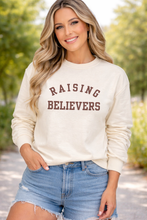 Load image into Gallery viewer, Woman wearing a cream sweatshirt with 'RAISING BELIEVERS' text, standing outdoors.