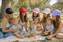 Load image into Gallery viewer, Group of women and children sitting on a blanket in a park, wearing branded hats.
