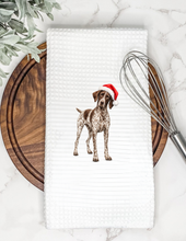 Load image into Gallery viewer, A white waffle-weave kitchen towel featuring a printed illustration of a brown and white spotted dog wearing a red Santa hat, styled on a marble surface with a wooden board, greenery, and a metal whisk.
