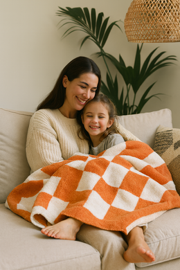 Woman and child sitting on a couch under an orange and white checkered blanket, with plants in the background.