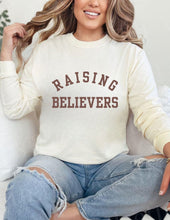 Load image into Gallery viewer, Person wearing a cream sweatshirt with 'RAISING BELIEVERS' text, sitting on a couch.
