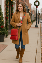 Load image into Gallery viewer, Woman in a tan coat with a red bag walking on a sidewalk decorated for Christmas.
