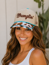 Load image into Gallery viewer, Woman wearing a beige cap with 'Wranglin'' text and a cow print design, smiling outdoors.

